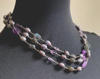 May include: A multi-strand necklace featuring a combination of grey pearls, translucent purple beads, and iridescent grey stones. The beads are strung on a thin, light-colored cord, creating a layered effect with varied textures and shapes.