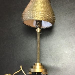 May include: A table lamp with a metallic gold-toned, woven-textured shade and a slender, braided-style stem. The base is a tiered, circular metallic gold-toned structure. A white electrical cord with a standard plug extends from the base.
