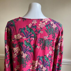 May include: A long-sleeved, pink blouse with a vibrant floral and bird print. The design features green parrots, white and pink flowers, and blue accents. The blouse is displayed on a white mannequin.