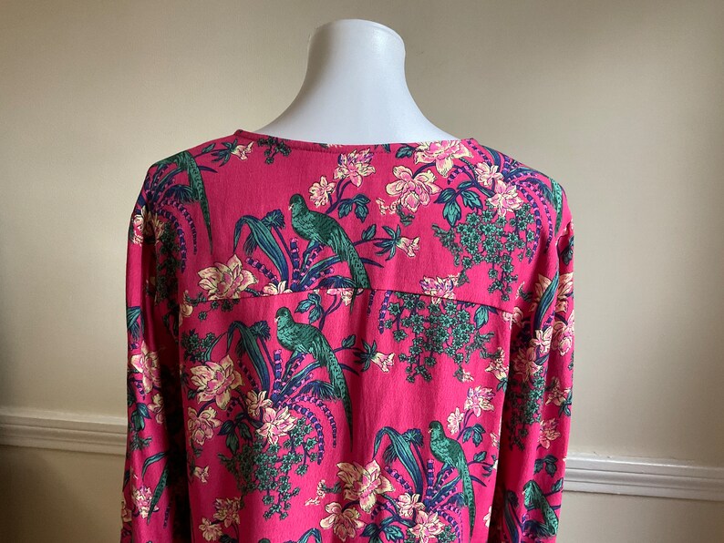 May include: A long-sleeved, pink blouse with a vibrant floral and bird print. The design features green parrots, white and pink flowers, and blue accents. The blouse is displayed on a white mannequin.