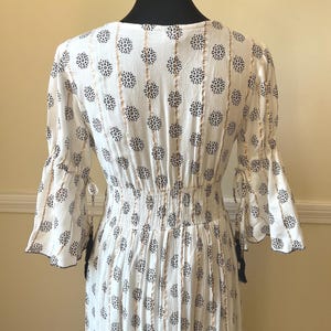 May include: White dress with a floral pattern and gold vertical stripes. The dress has flared sleeves with tie details and a cinched waist. The fabric has a textured appearance, and the dress is displayed on a black mannequin.