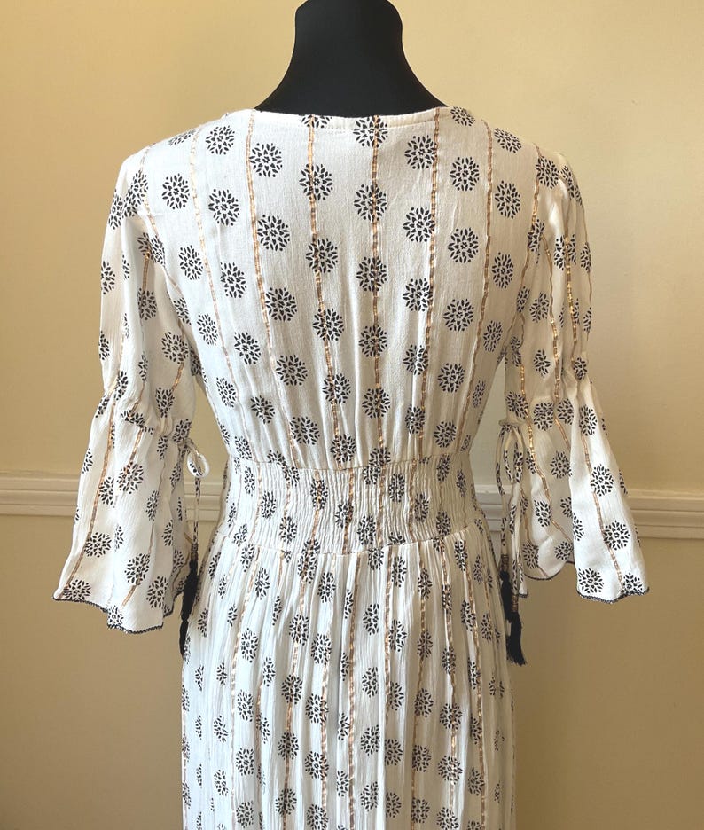 May include: White dress with a floral pattern and gold vertical stripes. The dress has flared sleeves with tie details and a cinched waist. The fabric has a textured appearance, and the dress is displayed on a black mannequin.
