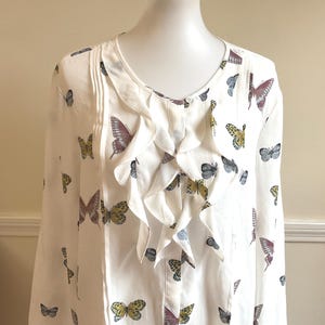 May include: A white long-sleeve blouse featuring a repeating pattern of butterflies in shades of yellow, purple, and grey. The garment has a round neckline and a vertical ruffled placket down the center front, with pleats on either side of the chest.