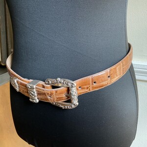 Puede incluir: Un cintur&oacute;n de color tostado con un patr&oacute;n texturizado de relieve de cocodrilo y una hebilla de metal plateado con intrincados grabados y detalles trenzados. El cintur&oacute;n incluye una trabilla de metal plateado a juego y una punta.