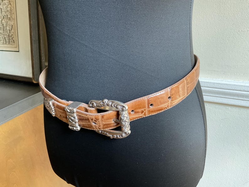 Puede incluir: Un cintur&oacute;n de color tostado con un patr&oacute;n texturizado de relieve de cocodrilo y una hebilla de metal plateado con intrincados grabados y detalles trenzados. El cintur&oacute;n incluye una trabilla de metal plateado a juego y una punta.