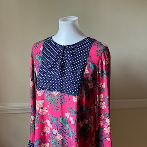 May include: A long-sleeved dress with a vibrant pink floral pattern featuring green birds and white flowers. The yoke is navy blue with white polka dots. The dress is displayed on a white mannequin.
