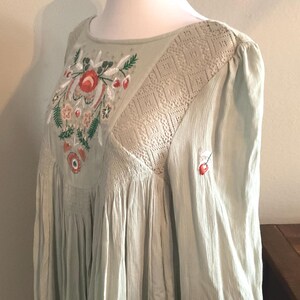 K&ouml;nnte beinhalten: Hellgr&uuml;nes Kleid mit floraler Stickerei auf dem Oberteil. Das Kleid hat einen runden Ausschnitt, lange &Auml;rmel und eine flie&szlig;ende Silhouette. Die Stickerei umfasst rote, orangefarbene und gr&uuml;ne Blumenmuster. Das Kleid hat einen strukturierten Stoff und eine Spitzeneinsatz.