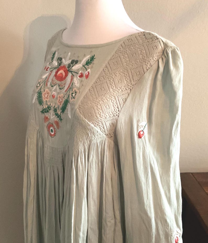 K&ouml;nnte beinhalten: Hellgr&uuml;nes Kleid mit floraler Stickerei auf dem Oberteil. Das Kleid hat einen runden Ausschnitt, lange &Auml;rmel und eine flie&szlig;ende Silhouette. Die Stickerei umfasst rote, orangefarbene und gr&uuml;ne Blumenmuster. Das Kleid hat einen strukturierten Stoff und eine Spitzeneinsatz.