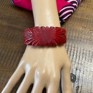 May include: A deep red bangle bracelet featuring a carved, textured surface with radiating leaf-like patterns. The wide, solid-colored cuff has a smooth, polished finish and a scalloped edge design that wraps around the wrist area.