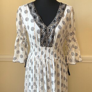 May include: White dress with a V-neck and three-quarter sleeves. The dress features a black and white floral pattern, gold vertical stripes, and decorative trim around the neckline. The dress is displayed on a black mannequin.
