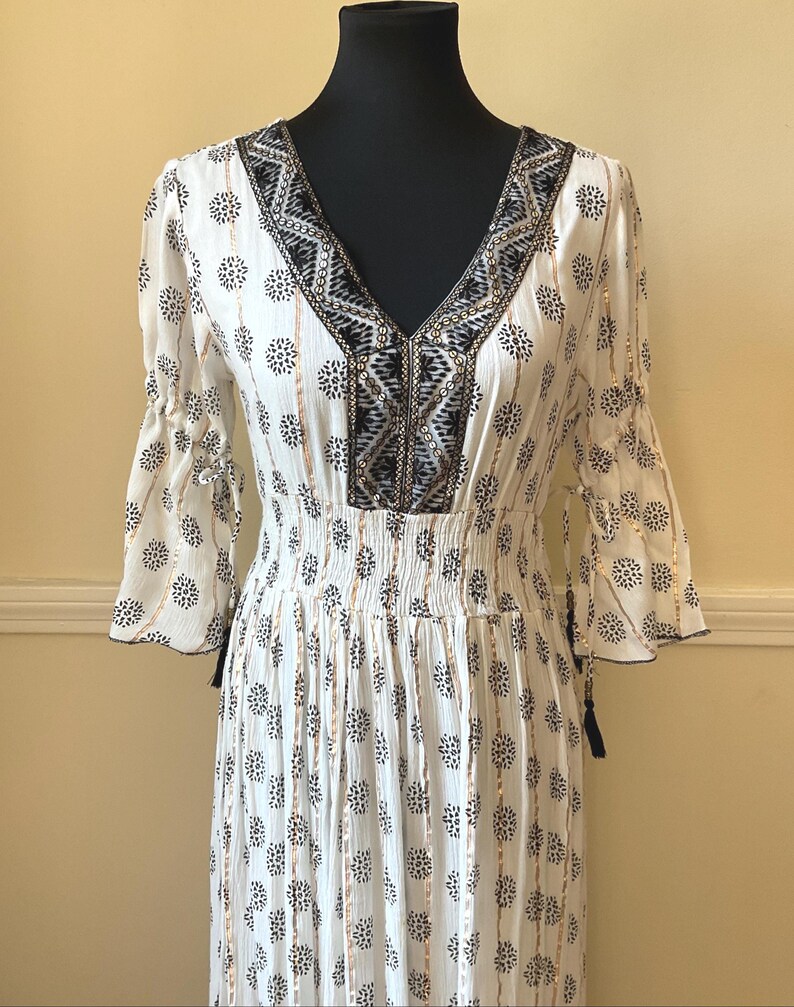 May include: White dress with a V-neck and three-quarter sleeves. The dress features a black and white floral pattern, gold vertical stripes, and decorative trim around the neckline. The dress is displayed on a black mannequin.