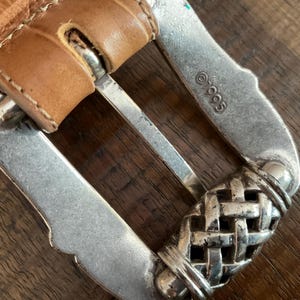 Puede incluir: Un cintur&oacute;n de cuero color canela con una superficie texturizada y en relieve y una hebilla de metal plateado. La hebilla presenta un patr&oacute;n de tejido de cesta en el extremo y est&aacute; estampada con el texto 1995. La correa se asegura con un perno de metal.