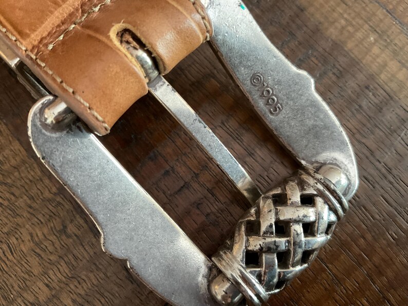 Puede incluir: Un cintur&oacute;n de cuero color canela con una superficie texturizada y en relieve y una hebilla de metal plateado. La hebilla presenta un patr&oacute;n de tejido de cesta en el extremo y est&aacute; estampada con el texto 1995. La correa se asegura con un perno de metal.