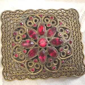May include: A square brass brooch featuring an intricate filigree border and a central floral motif. The design is embellished with numerous faceted red glass stones arranged in a starburst pattern, accented by smaller red stones throughout.