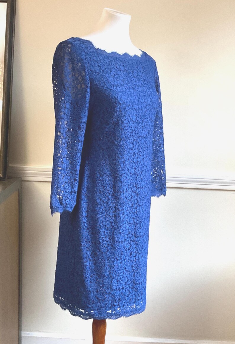 May include: A cobalt blue lace dress with long sleeves and a scalloped neckline. The knee-length dress is displayed on a mannequin. The dress has a floral lace pattern and a scalloped hemline. The dress is a vintage style.