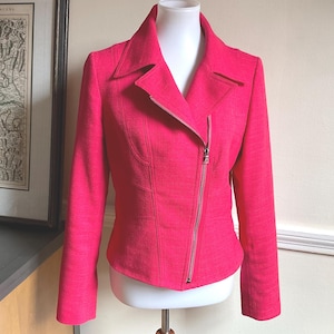 May include: A bright pink jacket with a diagonal zipper closure and a notched collar. The jacket is displayed on a mannequin and has long sleeves. The fabric has a subtle textured appearance.