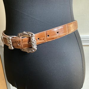 Puede incluir: Un cintur&oacute;n de cuero color canela con un acabado brillante y texturizado de imitaci&oacute;n cocodrilo. El cintur&oacute;n cuenta con una hebilla grande de metal plateado con intrincados adornos y un dise&ntilde;o central trenzado, con una trabilla a juego.