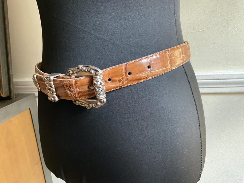 Puede incluir: Un cintur&oacute;n de cuero color canela con un acabado brillante y texturizado de imitaci&oacute;n cocodrilo. El cintur&oacute;n cuenta con una hebilla grande de metal plateado con intrincados adornos y un dise&ntilde;o central trenzado, con una trabilla a juego.