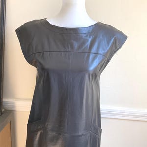 May include: A grey leather dress with a round neckline and short sleeves. The dress has two front pockets and a tailored fit. The dress is displayed on a white mannequin. The dress is a classic, minimalist design.