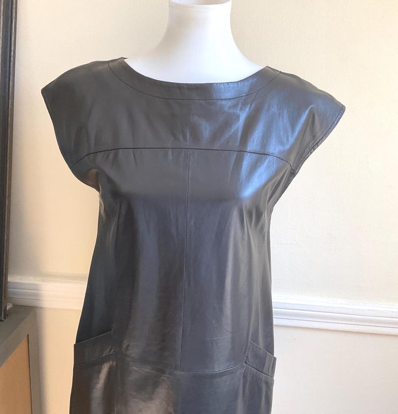 May include: A grey leather dress with a round neckline and short sleeves. The dress has two front pockets and a tailored fit. The dress is displayed on a white mannequin. The dress is a classic, minimalist design.