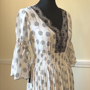 May include: White dress with a V-neck and bell sleeves. The dress features a black floral pattern and gold vertical stripes. The neckline and sleeves are trimmed with black embroidery. The waistline is elasticized.