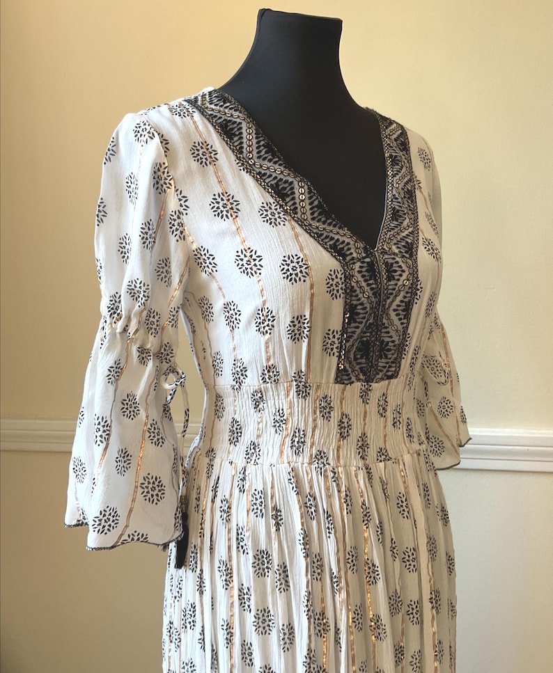 May include: White dress with a V-neck and bell sleeves. The dress features a black floral pattern and gold vertical stripes. The neckline and sleeves are trimmed with black embroidery. The waistline is elasticized.