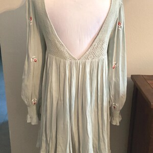 K&ouml;nnte beinhalten: Hellgr&uuml;nes Kleid mit langen &Auml;rmeln und tiefem R&uuml;ckenausschnitt. Das Kleid hat gestickte rote Blumen auf den &Auml;rmeln und ein strukturiertes R&uuml;ckenteil. Die &Auml;rmel haben ger&uuml;schte Manschetten, und der Rock ist gerafft.