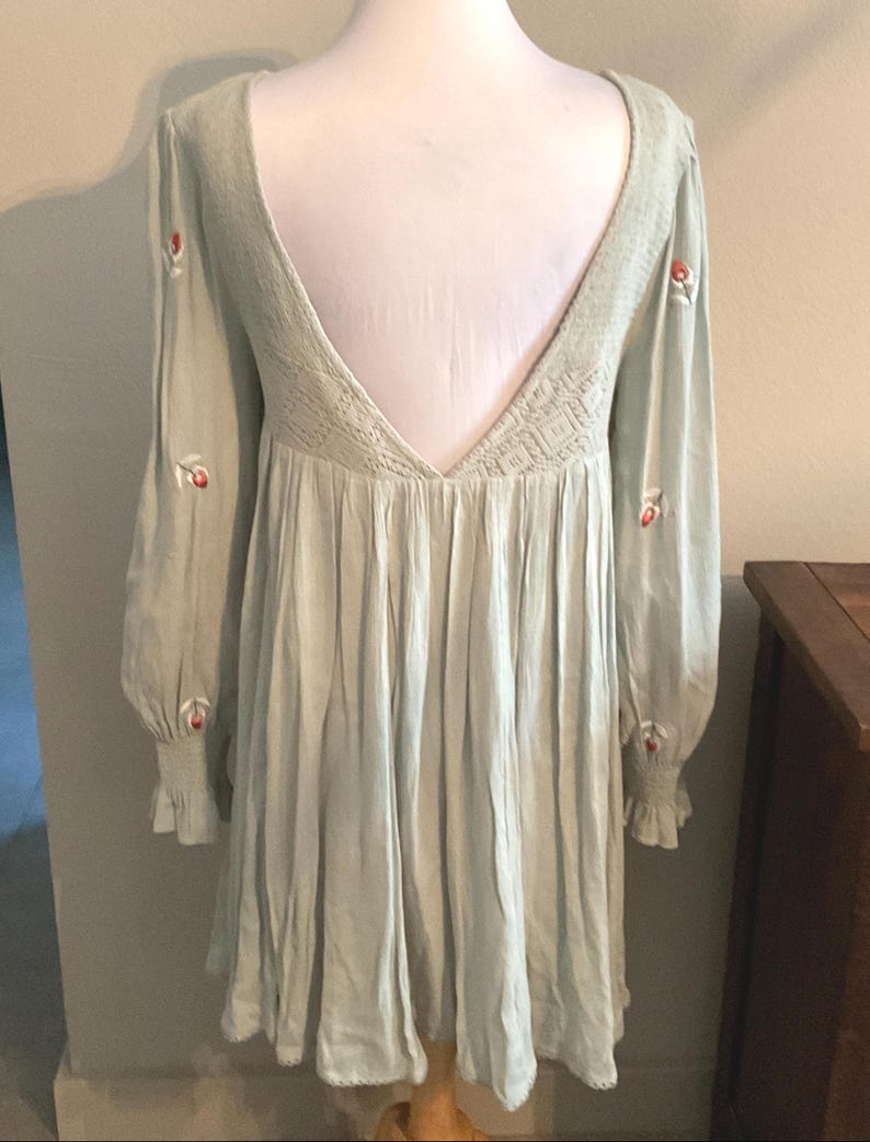 K&ouml;nnte beinhalten: Hellgr&uuml;nes Kleid mit langen &Auml;rmeln und tiefem R&uuml;ckenausschnitt. Das Kleid hat gestickte rote Blumen auf den &Auml;rmeln und ein strukturiertes R&uuml;ckenteil. Die &Auml;rmel haben ger&uuml;schte Manschetten, und der Rock ist gerafft.