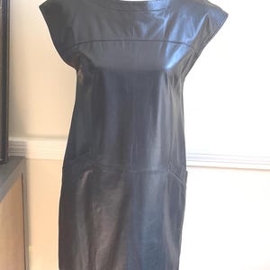 May include: A black leather dress with a round neckline and short sleeves. The dress has a straight silhouette and two front pockets. The dress is displayed on a mannequin.