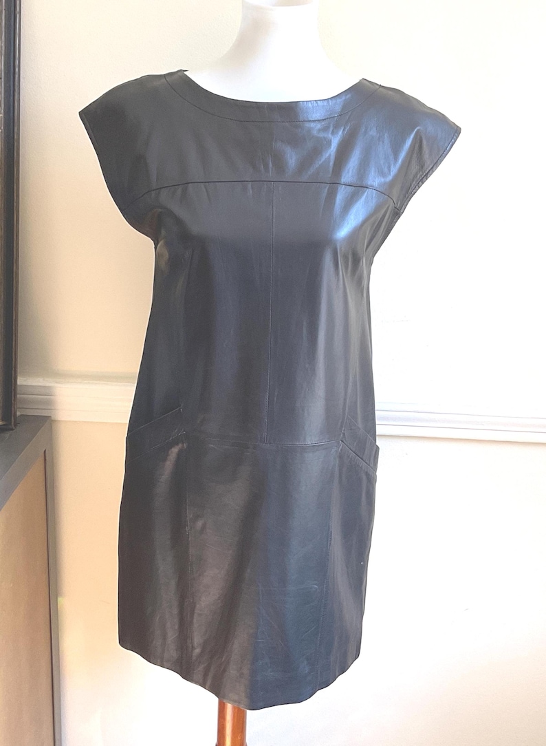 May include: A black leather dress with a round neckline and short sleeves. The dress has a straight silhouette and two front pockets. The dress is displayed on a mannequin.