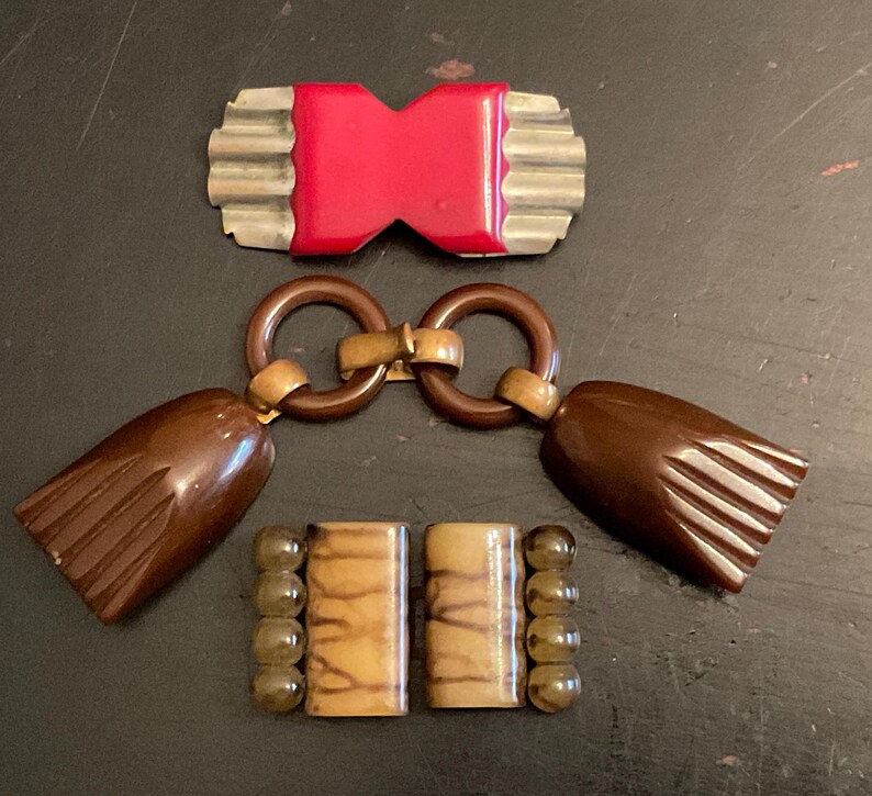 May include: Three vintage plastic and metal belt buckles. One is red and has a wavy metal frame. Another is brown with a tassel and a gold metal connector. The last is brown with a striped pattern and gold beads.