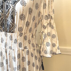May include: White dress with a floral pattern and gold stripes. The dress features a V-neckline with black embroidery, bell sleeves, and a gathered waist. A black tassel hangs from the sleeve.