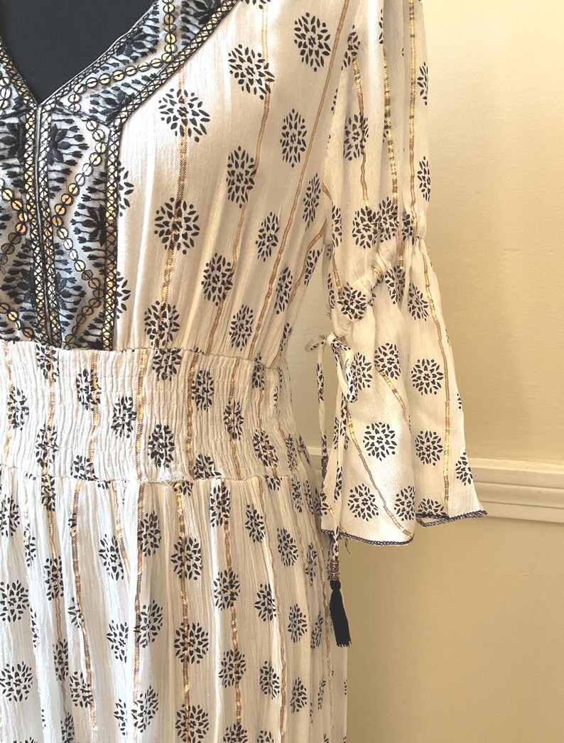 May include: White dress with a floral pattern and gold stripes. The dress features a V-neckline with black embroidery, bell sleeves, and a gathered waist. A black tassel hangs from the sleeve.