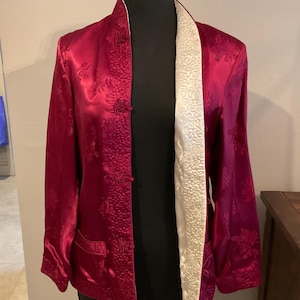 May include: A red satin jacket with a mandarin collar and floral embroidery. The jacket has a cream-colored lining and decorative frog closures. The jacket is displayed on a black mannequin.