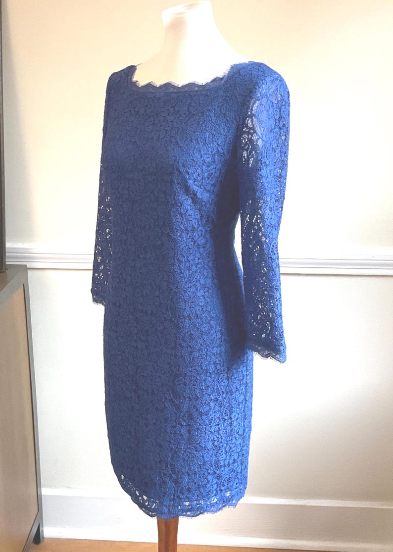May include: A navy blue lace dress with a scalloped neckline and three-quarter sleeves. The dress is displayed on a mannequin. The dress is knee-length and has a fitted silhouette.