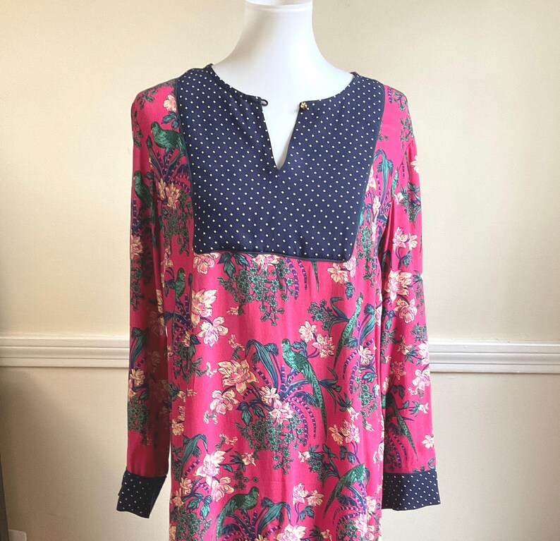 May include: A long-sleeved dress with a vibrant pink floral pattern featuring green birds and white flowers. The neckline and cuffs are navy blue with white polka dots. The neckline has a small gold-colored button.