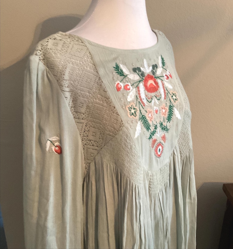 K&ouml;nnte beinhalten: Hellgr&uuml;nes Kleid mit floraler Stickerei. Das Kleid hat einen runden Ausschnitt, lange &Auml;rmel und ein detailliertes Stickdesign auf dem Oberteil. Die Stickerei umfasst rote, orangefarbene, gr&uuml;ne und wei&szlig;e florale Elemente. Das Kleid hat einen strukturierten Stoff und eine lockere Passform.