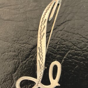 May include: A silver-toned metal brooch shaped like the cursive letter L. The elongated vertical stroke features a decorative etched pattern of small, repeating leaf-like shapes along its edge, while the base forms a smooth, rounded loop.