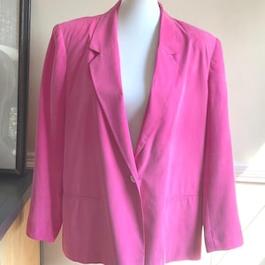 May include: A bright pink blazer displayed on a white mannequin. The blazer has a single button closure, notched lapels, and two front pockets. The jacket is made of a smooth fabric and has long sleeves.