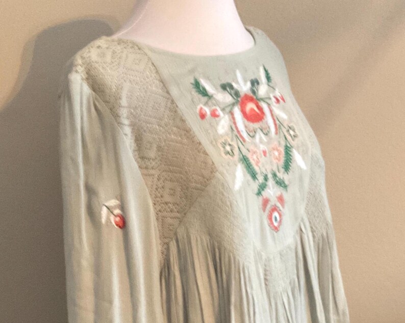 K&ouml;nnte beinhalten: Eine hellgr&uuml;ne, bestickte Tunika mit rundem Ausschnitt. Die Tunika hat florale Stickereien in Rot, Wei&szlig; und Gr&uuml;n auf der Vorderseite. Die &Auml;rmel sind lang und die Schultern haben Spitzendetails. Auf dem &Auml;rmel ist eine kleine Kirsche gestickt.