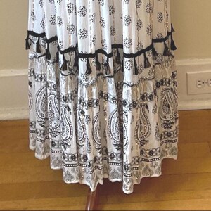 May include: White and black patterned dress with a paisley and floral design. The dress features black tassels and metallic accents. The skirt has multiple tiers and a flowing silhouette, creating a bohemian aesthetic.