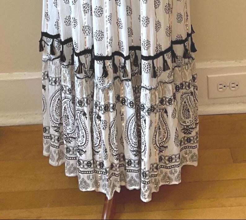 May include: White and black patterned dress with a paisley and floral design. The dress features black tassels and metallic accents. The skirt has multiple tiers and a flowing silhouette, creating a bohemian aesthetic.