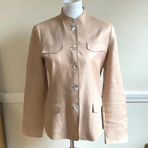 May include: A light tan jacket with a mandarin collar and four pearlescent buttons. The jacket has two chest pockets and two lower pockets. The stitching is visible around the pockets and seams. The jacket is displayed on a mannequin.