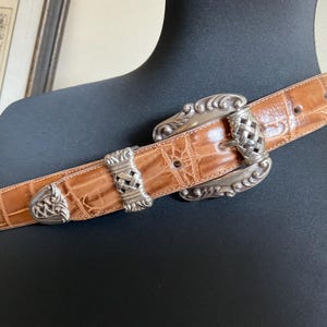 Puede incluir: Un cintur&oacute;n de color tostado con un patr&oacute;n texturizado de relieve de cocodrilo, que presenta una hebilla grande y ornamentada de metal plateado y detalles de metal plateado a juego en la correa, incluyendo una trabilla y una punta.