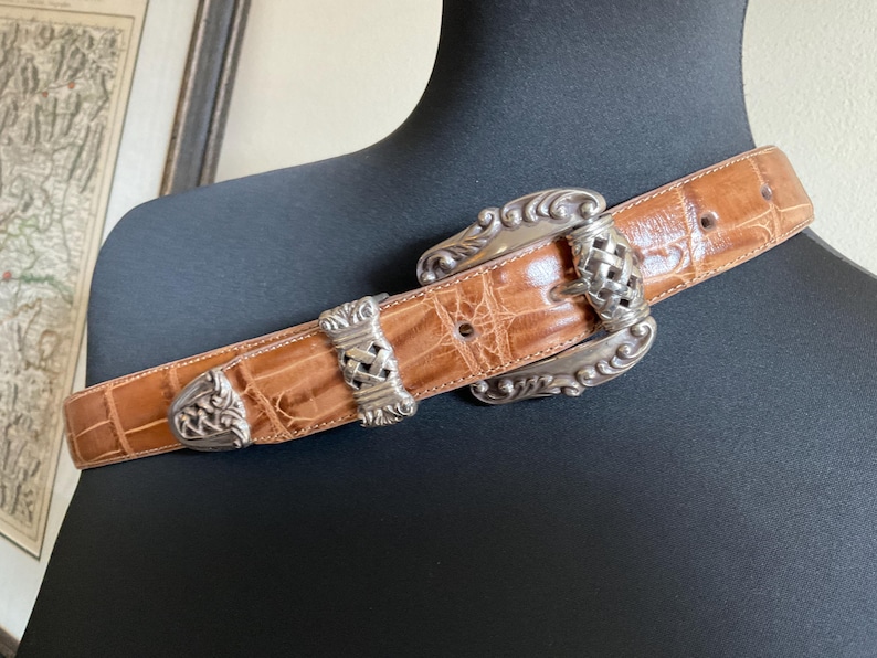 Puede incluir: Un cintur&oacute;n de color tostado con un patr&oacute;n texturizado de relieve de cocodrilo, que presenta una hebilla grande y ornamentada de metal plateado y detalles de metal plateado a juego en la correa, incluyendo una trabilla y una punta.