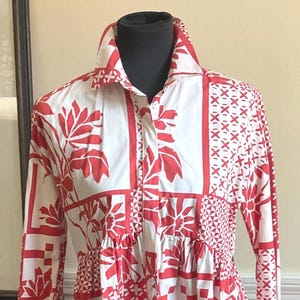 May include: A long-sleeved button-down shirt featuring a white base with a vibrant red patchwork-style print. The pattern consists of alternating blocks of floral motifs, geometric shapes, and cross-hatch designs. It has a classic collar.