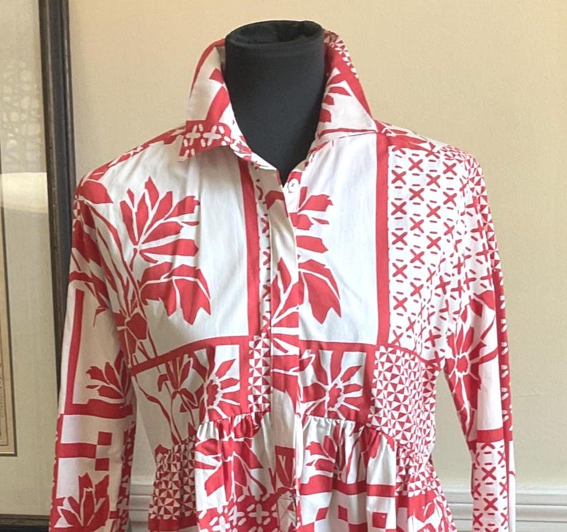 May include: A long-sleeved button-down shirt featuring a white base with a vibrant red patchwork-style print. The pattern consists of alternating blocks of floral motifs, geometric shapes, and cross-hatch designs. It has a classic collar.