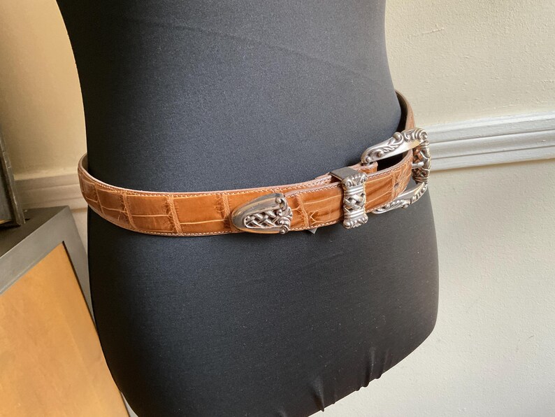 Puede incluir: Un cintur&oacute;n de color tostado con un patr&oacute;n de relieve de cocodrilo que presenta una hebilla de metal plateado, una punta de metal plateado a juego y una trabilla decorativa de metal plateado, con intrincados detalles grabados.