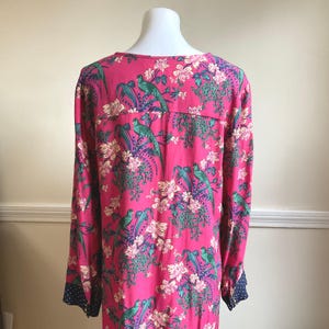 May include: A long-sleeved, pink dress featuring a vibrant floral and bird pattern. The design includes green parrots, white flowers, and blue accents. The sleeves have a navy blue cuff with white polka dots. The dress is displayed on a mannequin.
