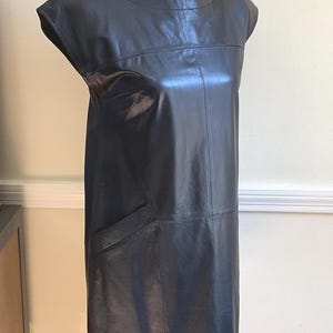 May include: A black leather dress with a round neckline and short sleeves. The dress has a straight silhouette and a single pocket on the front. The dress is displayed on a mannequin.